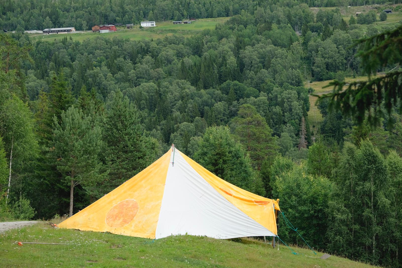 Stort oransje og hvitt stripete telt på grønn eng med utsikt over skogkledt dal og røde gårdsbygninger